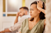 A woman with a serious expression sits in the foreground, resting one hand on her head. A man in the background sits blurred, looking in the opposite direction. Both appear to be in a tense or thoughtful moment indoors.