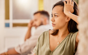 A woman with a serious expression sits in the foreground, resting one hand on her head. A man in the background sits blurred, looking in the opposite direction. Both appear to be in a tense or thoughtful moment indoors.