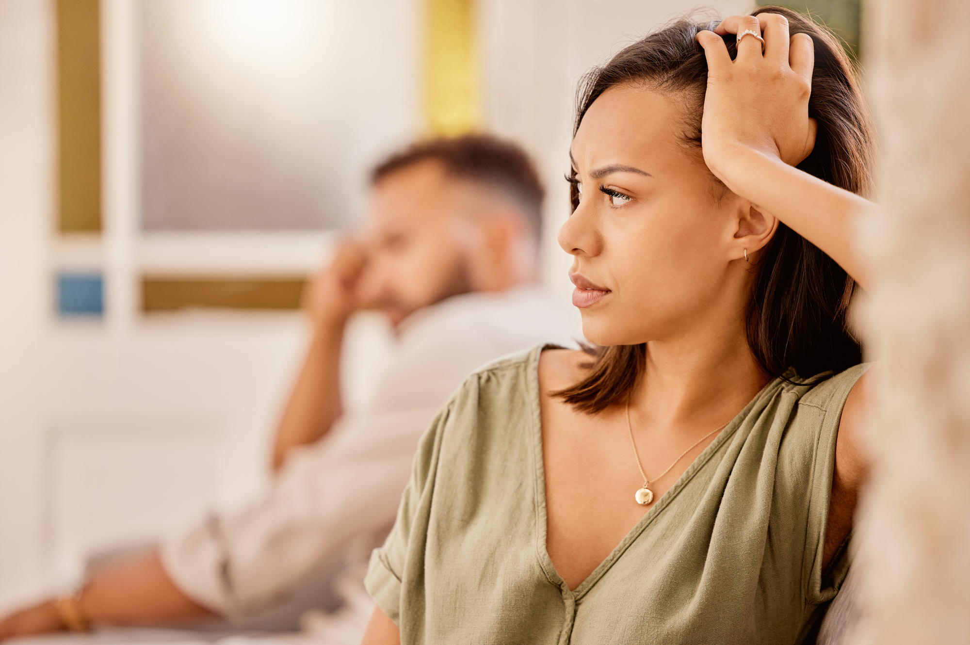 A woman with a serious expression sits in the foreground, resting one hand on her head. A man in the background sits blurred, looking in the opposite direction. Both appear to be in a tense or thoughtful moment indoors.