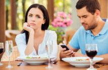 A woman and a man are sitting at a dining table with plates of salad and glasses of water and red wine. The woman looks bored and rests her chin on her hand while the man is busy looking at his phone. The background features blurred outdoor greenery and flowers.