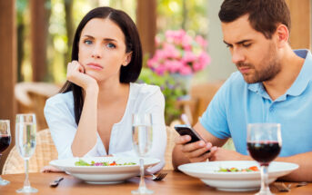 A woman and a man are sitting at a dining table with plates of salad and glasses of water and red wine. The woman looks bored and rests her chin on her hand while the man is busy looking at his phone. The background features blurred outdoor greenery and flowers.