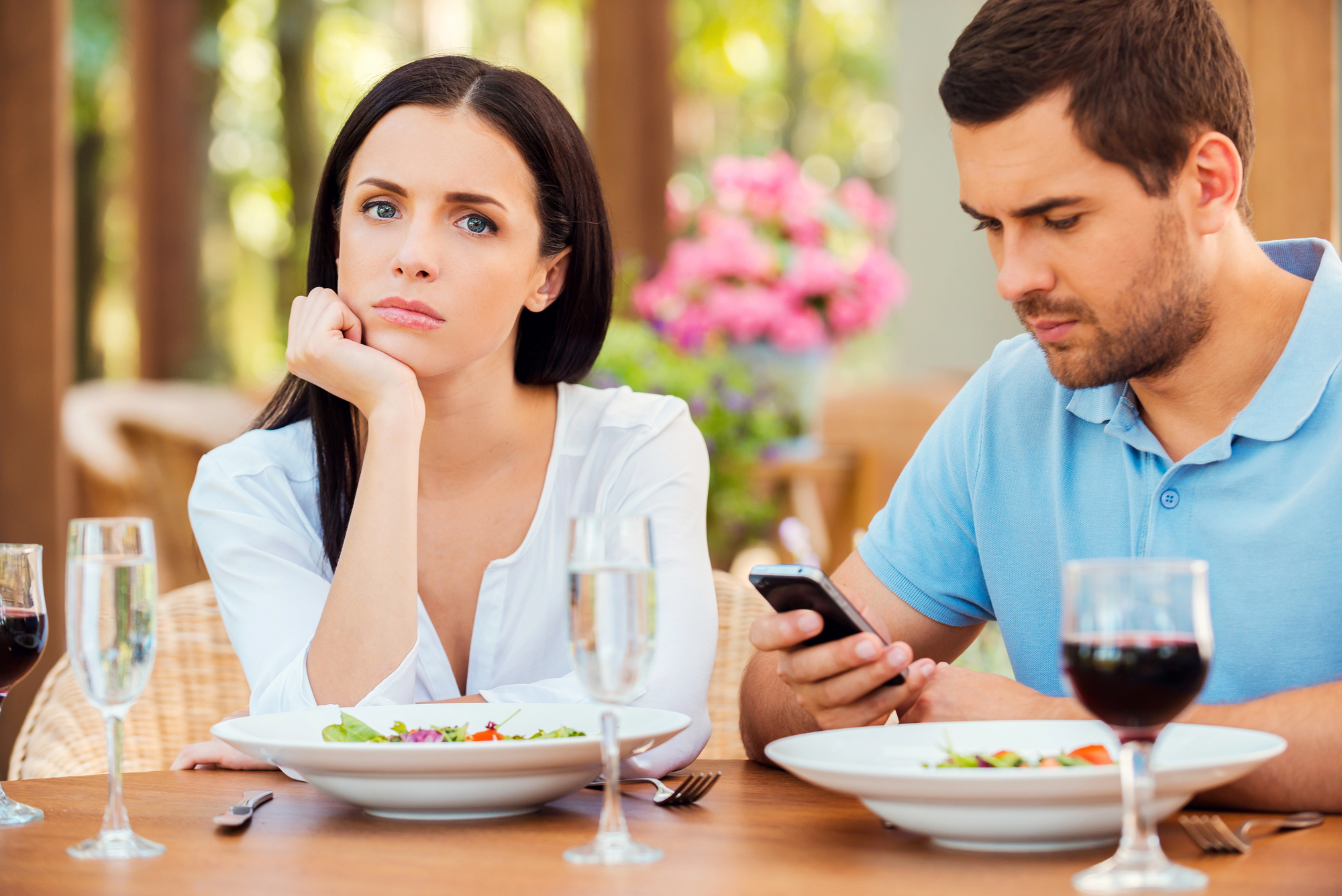 A woman and a man are sitting at a dining table with plates of salad and glasses of water and red wine. The woman looks bored and rests her chin on her hand while the man is busy looking at his phone. The background features blurred outdoor greenery and flowers.