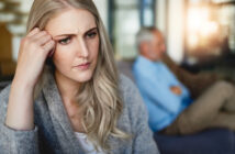 A young woman with long blonde hair looks worried while sitting indoors, resting her hand on her temple. In the blurred background, an older man with gray hair and a beard sits with his arms crossed, also appearing concerned.