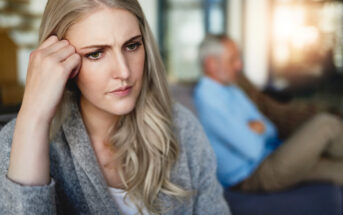 A young woman with long blonde hair looks worried while sitting indoors, resting her hand on her temple. In the blurred background, an older man with gray hair and a beard sits with his arms crossed, also appearing concerned.