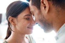 A close-up image of a couple with their foreheads touching and eyes closed, sharing an intimate and serene moment. The woman has her hair tied back and is wearing a delicate earring, while the man has short hair and facial hair. Both appear content and peaceful.