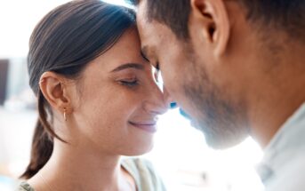 A close-up image of a couple with their foreheads touching and eyes closed, sharing an intimate and serene moment. The woman has her hair tied back and is wearing a delicate earring, while the man has short hair and facial hair. Both appear content and peaceful.