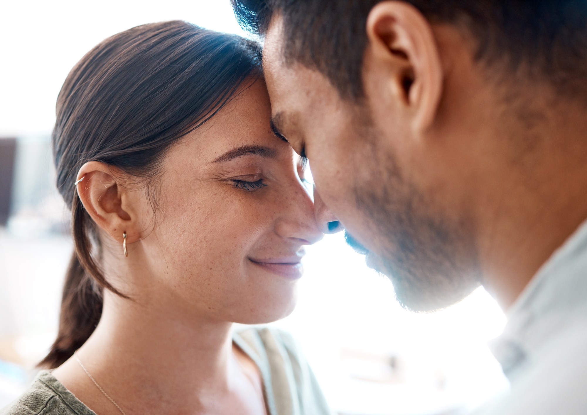 A close-up image of a couple with their foreheads touching and eyes closed, sharing an intimate and serene moment. The woman has her hair tied back and is wearing a delicate earring, while the man has short hair and facial hair. Both appear content and peaceful.