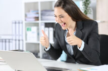 A woman in a business suit is sitting at a desk in an office, looking at her laptop with excitement. She has a big smile and is giving two thumbs up. The background features office shelves and plants.