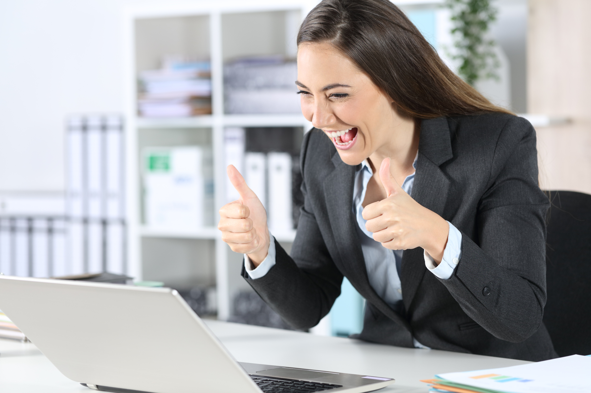 A woman in a business suit is sitting at a desk in an office, looking at her laptop with excitement. She has a big smile and is giving two thumbs up. The background features office shelves and plants.