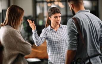 A woman in a blue checkered shirt stands in an office, gesturing with her hand while speaking to a man and a woman. The woman's expression is serious, and the man and woman appear to be listening intently. There is a table in the background.