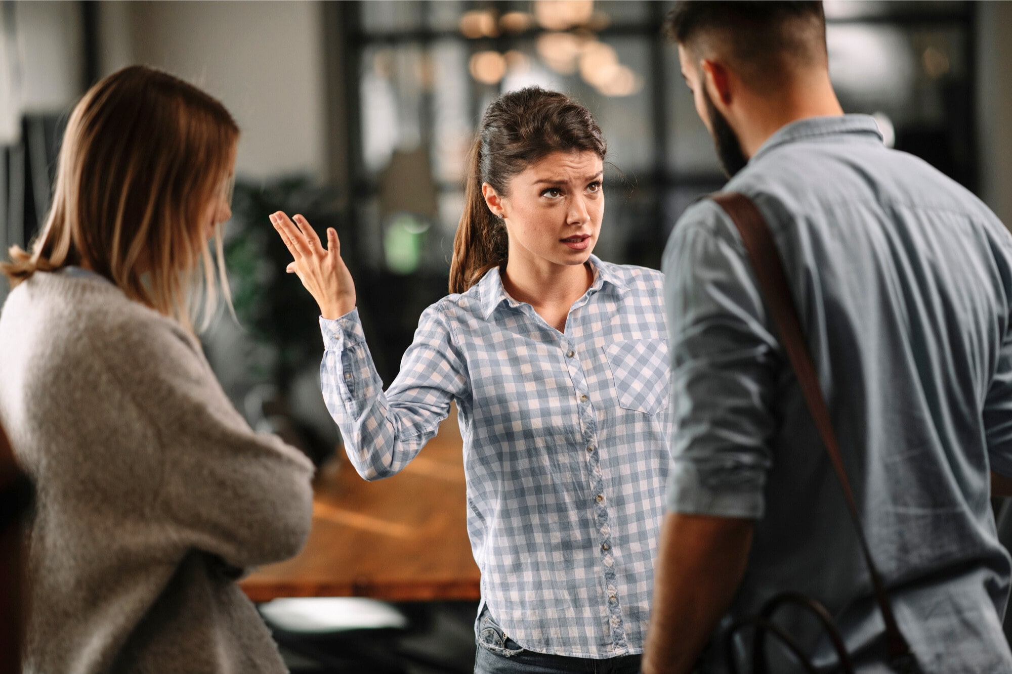 A woman in a blue checkered shirt stands in an office, gesturing with her hand while speaking to a man and a woman. The woman's expression is serious, and the man and woman appear to be listening intently. There is a table in the background.