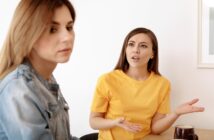 Two women sit at a table; the woman on the right, in a yellow shirt, appears to be speaking or expressing frustration, gesturing with her hands. The woman on the left, in a denim jacket, looks away with a neutral or indifferent expression.