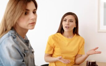 Two women sit at a table; the woman on the right, in a yellow shirt, appears to be speaking or expressing frustration, gesturing with her hands. The woman on the left, in a denim jacket, looks away with a neutral or indifferent expression.