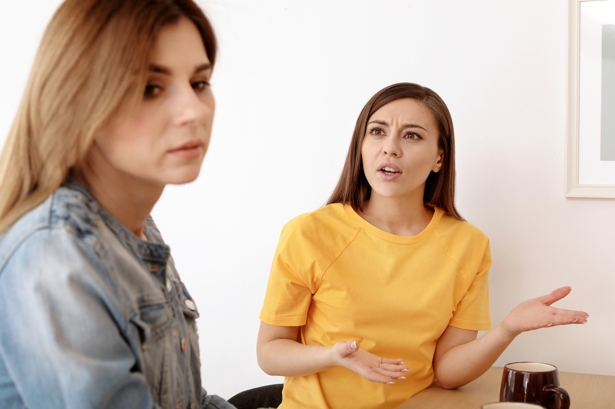 Two women sit at a table; the woman on the right, in a yellow shirt, appears to be speaking or expressing frustration, gesturing with her hands. The woman on the left, in a denim jacket, looks away with a neutral or indifferent expression.