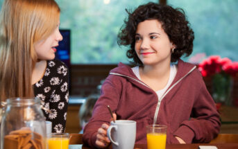 Two young girls sitting at a wooden table talking and smiling. One, with long blonde hair wearing a black flowered shirt, faces the other, with short curly dark hair in a maroon hoodie. On the table are glasses of orange juice, a mug, and various snacks.