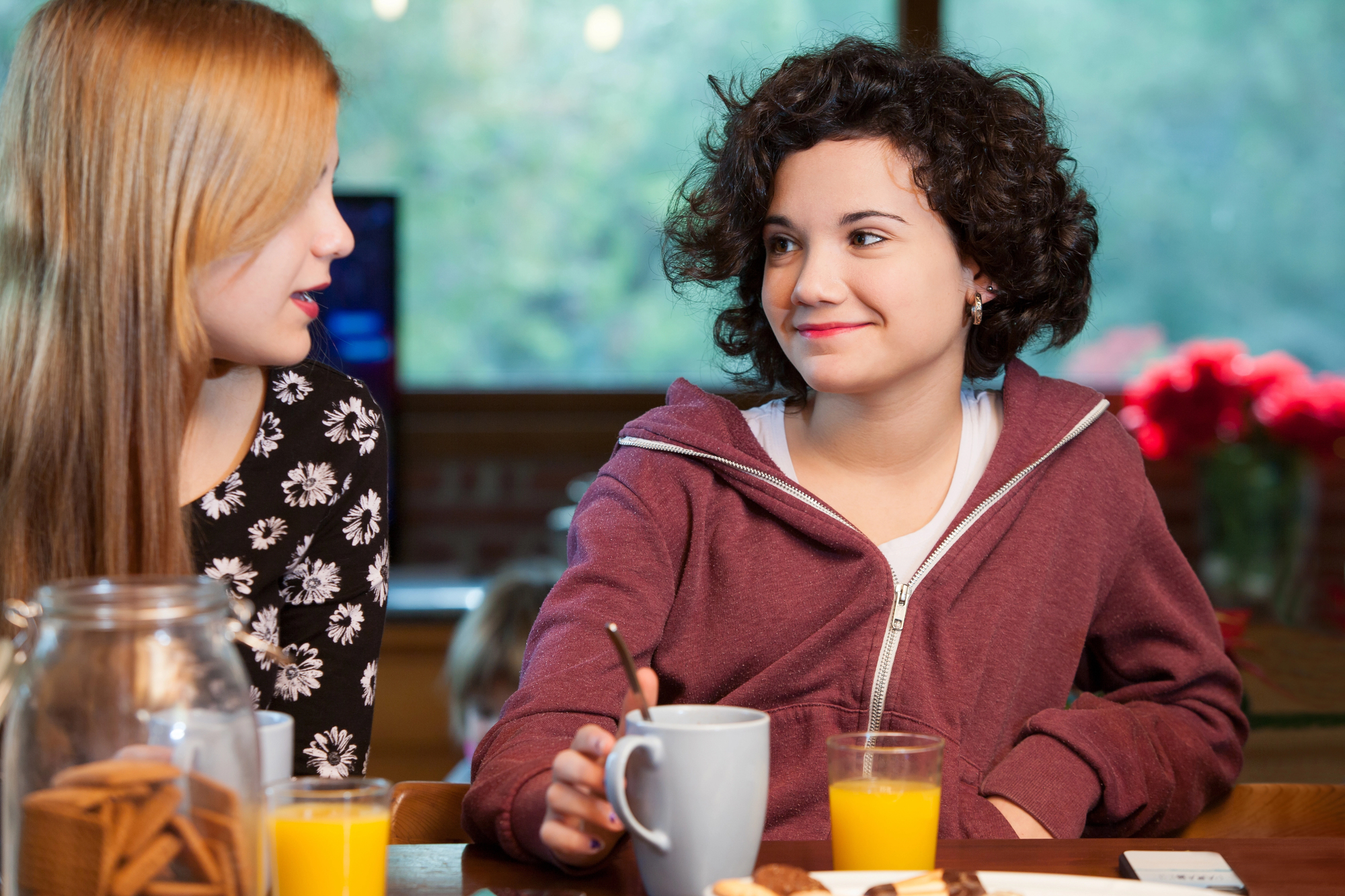 Two young girls sitting at a wooden table talking and smiling. One, with long blonde hair wearing a black flowered shirt, faces the other, with short curly dark hair in a maroon hoodie. On the table are glasses of orange juice, a mug, and various snacks.