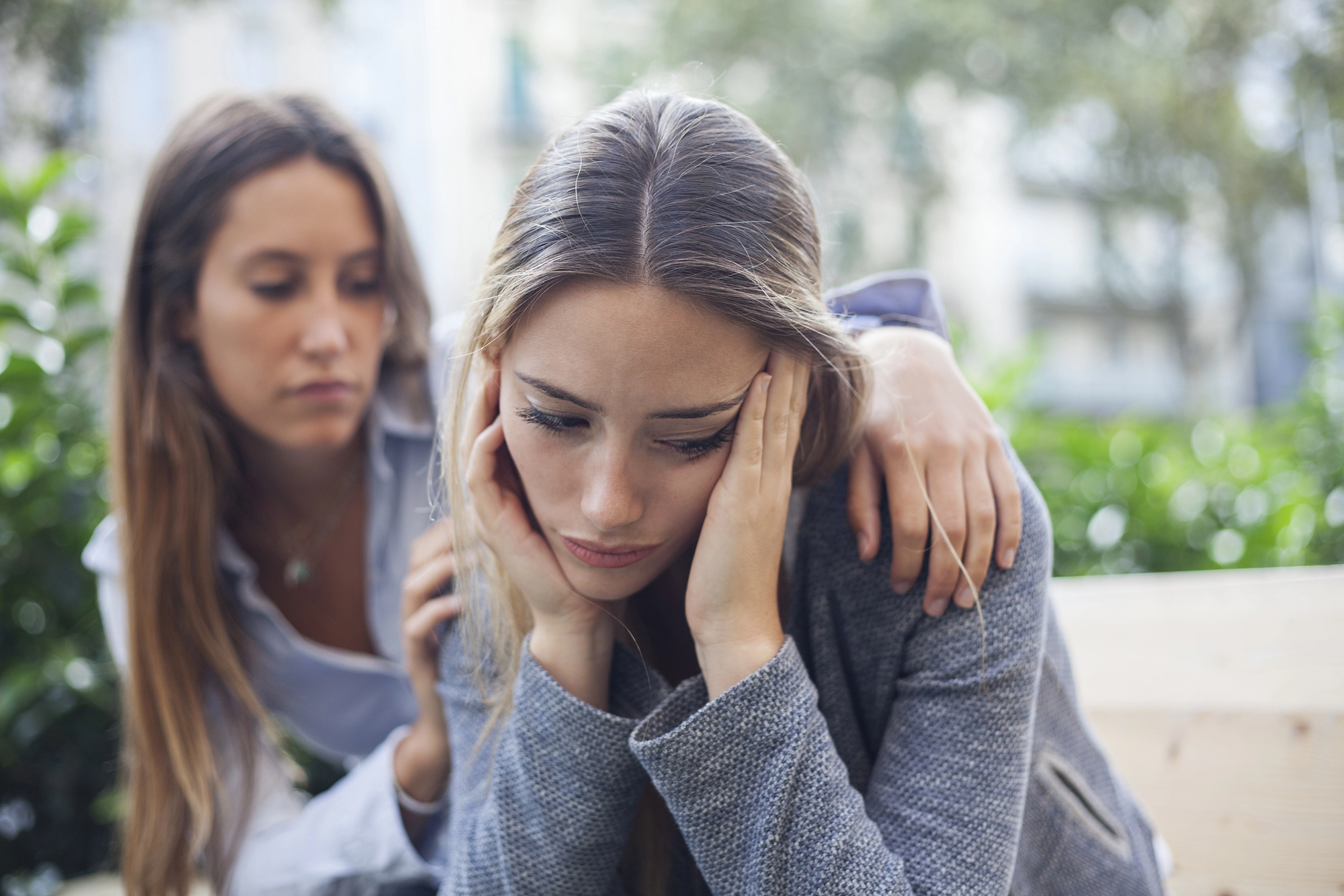 Two women are seated outdoors. The woman in the foreground appears distressed, holding her head in her hands. The woman behind her is offering comfort by placing a hand on her friend's shoulder. The background features blurred greenery and buildings.