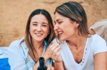 Two women sit closely together, sharing a moment of laughter and joy. One woman holds a beverage bottle, while the other smiles widely. Both wear casual clothing and jewelry, enjoying a relaxed outdoor setting with sandy ground visible in the background.