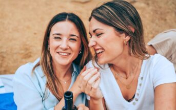 Two women sit closely together, sharing a moment of laughter and joy. One woman holds a beverage bottle, while the other smiles widely. Both wear casual clothing and jewelry, enjoying a relaxed outdoor setting with sandy ground visible in the background.