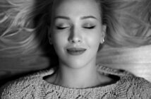 Black and white image of a woman with closed eyes, lying down with a peaceful expression. She has shoulder-length hair spread out around her and is wearing a textured knit sweater.