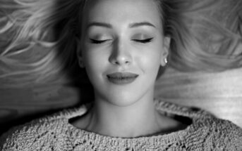 Black and white image of a woman with closed eyes, lying down with a peaceful expression. She has shoulder-length hair spread out around her and is wearing a textured knit sweater.