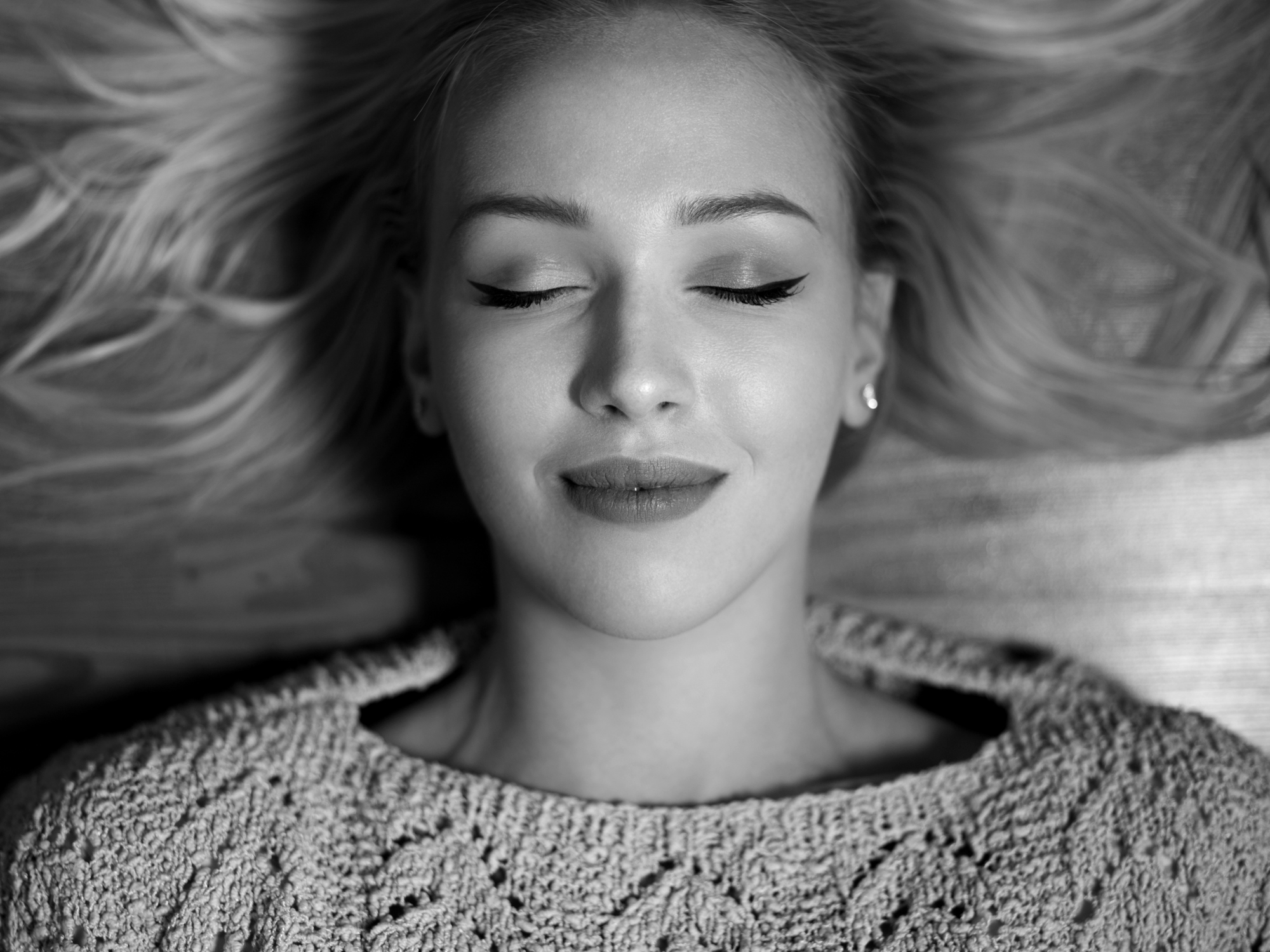 Black and white image of a woman with closed eyes, lying down with a peaceful expression. She has shoulder-length hair spread out around her and is wearing a textured knit sweater.