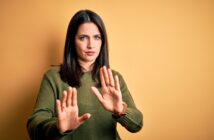 A woman with dark hair and a green sweater stands against a yellow background, holding up her hands in a "stop" gesture. She has a serious expression on her face.