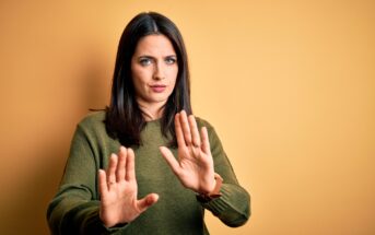 A woman with dark hair and a green sweater stands against a yellow background, holding up her hands in a "stop" gesture. She has a serious expression on her face.