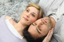 A woman with blonde hair and a man with dark hair lie next to each other on a fuzzy gray surface, gazing upwards. The woman is slightly smiling while gently touching the man's head, and both appear relaxed and content.