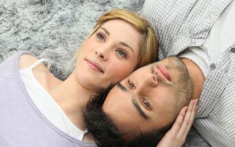 A woman with blonde hair and a man with dark hair lie next to each other on a fuzzy gray surface, gazing upwards. The woman is slightly smiling while gently touching the man's head, and both appear relaxed and content.