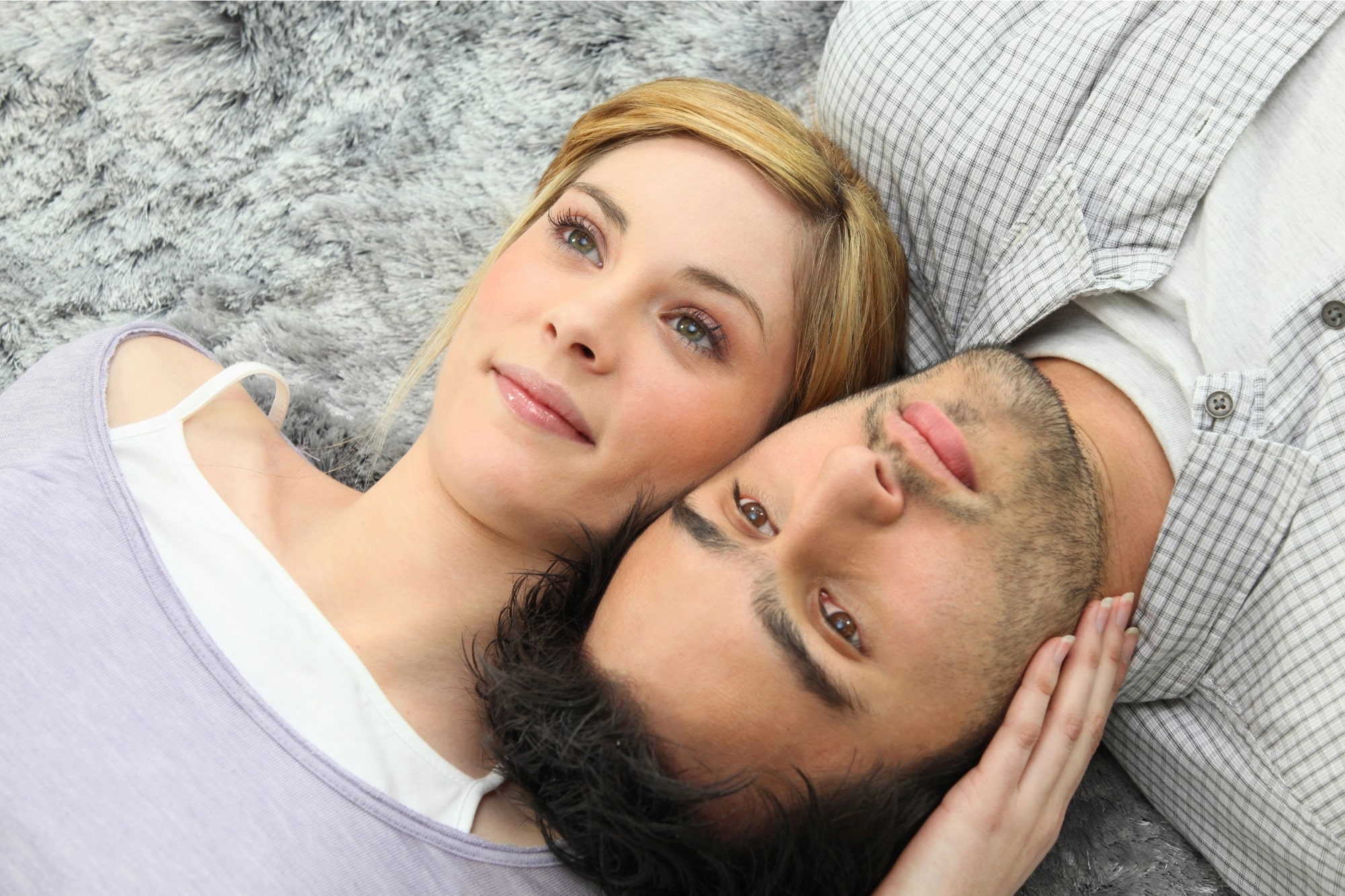 A woman with blonde hair and a man with dark hair lie next to each other on a fuzzy gray surface, gazing upwards. The woman is slightly smiling while gently touching the man's head, and both appear relaxed and content.