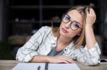 A young woman with long blonde hair and glasses sits at a desk, leaning her head on her hand. She is wearing a white checkered shirt over a gray top. In front of her is an open notebook with a pen. The background is blurred.