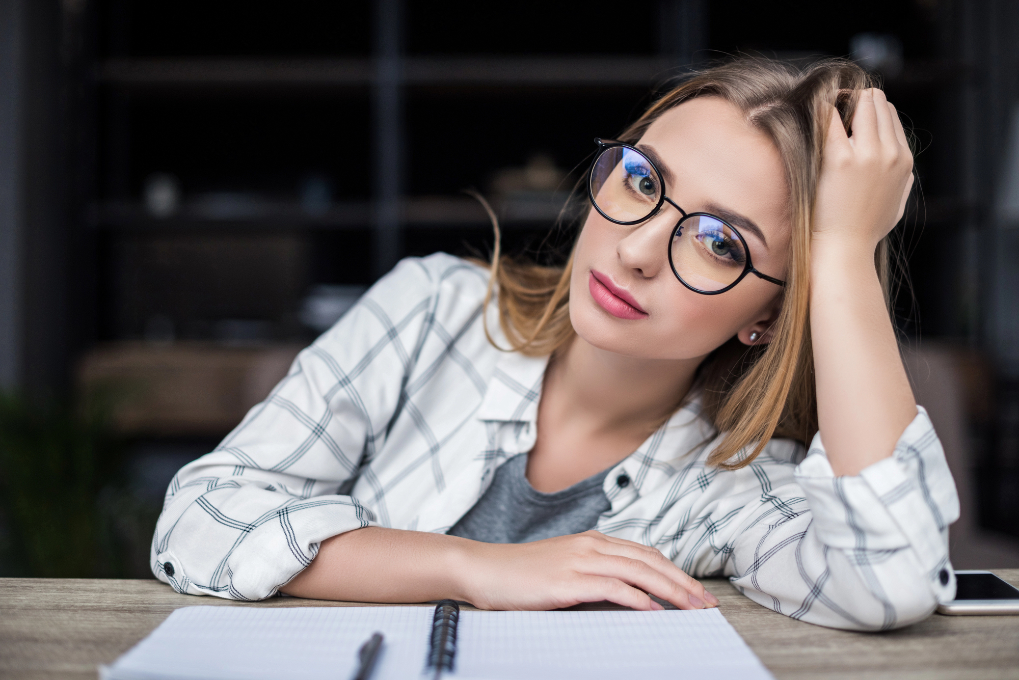 A young woman with long blonde hair and glasses sits at a desk, leaning her head on her hand. She is wearing a white checkered shirt over a gray top. In front of her is an open notebook with a pen. The background is blurred.