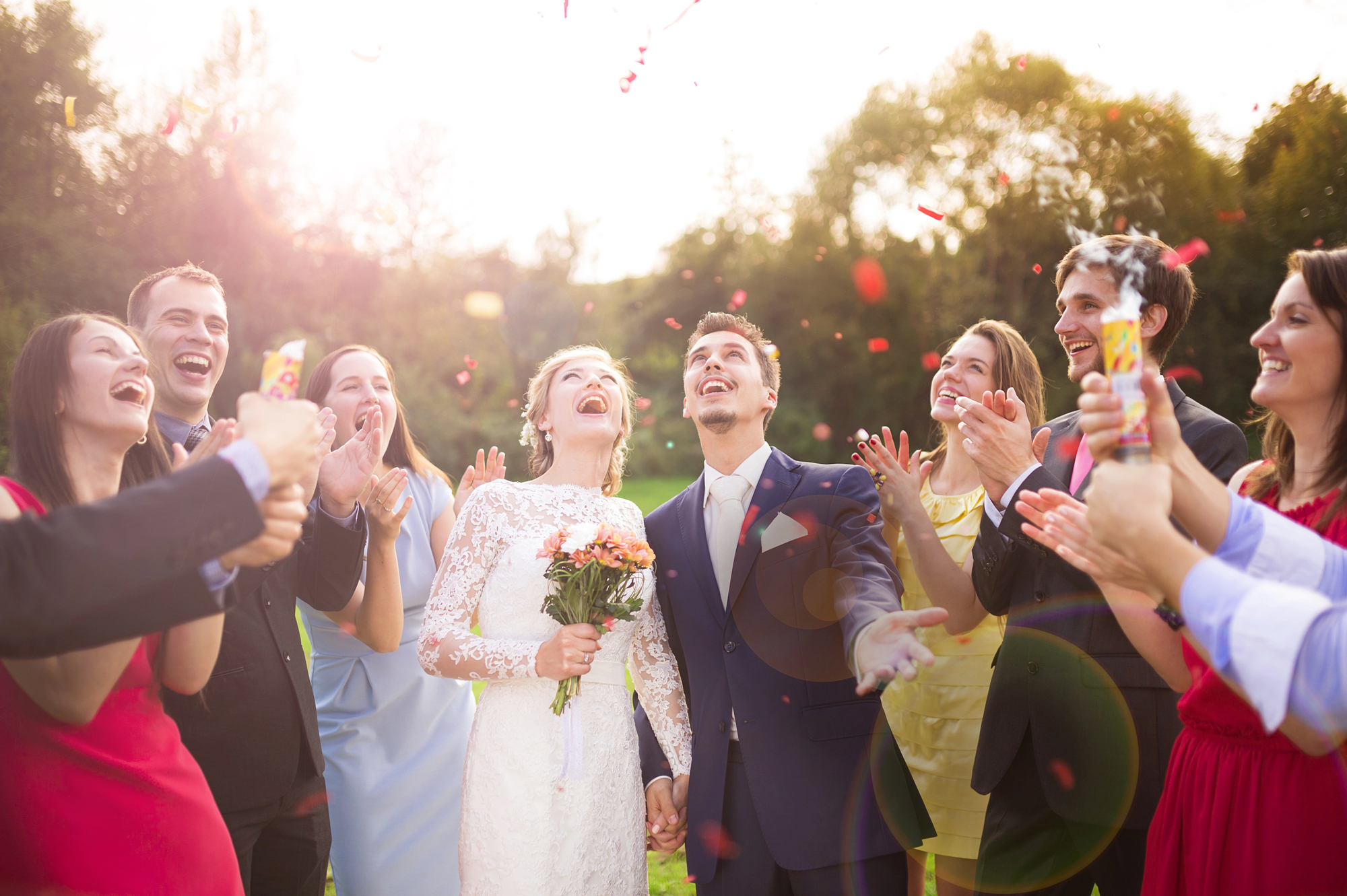 A bride and groom, both smiling, stand outdoors surrounded by cheering friends. The bride holds a bouquet of flowers, and colorful confetti is being thrown into the air. The joyful group, dressed in formal attire, celebrates on a sunny day with trees in the background.