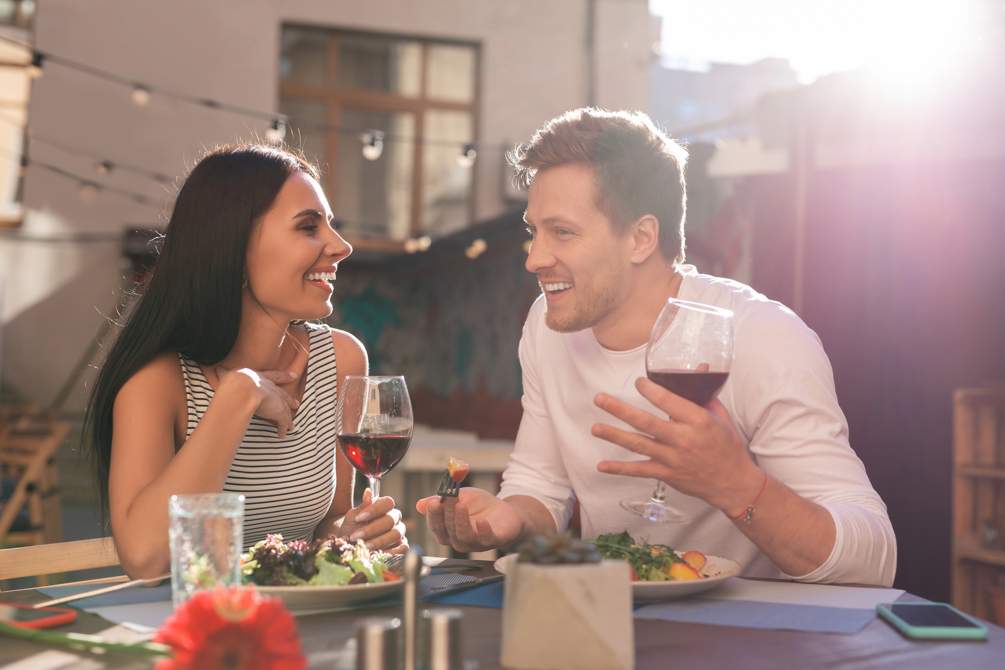 A man and woman sit at an outdoor table, smiling and conversing while holding glasses of red wine. The table is set with plates of food. String lights and a building facade are visible in the background, with sunlight streaming in.