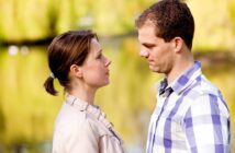 A man and a woman face each other, appearing to be in a serious conversation. They are outdoors near a body of water with greenery in the background. The woman has her hair tied back and wears a light jacket, while the man has short hair and wears a plaid shirt.