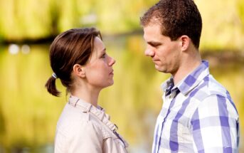 A man and a woman face each other, appearing to be in a serious conversation. They are outdoors near a body of water with greenery in the background. The woman has her hair tied back and wears a light jacket, while the man has short hair and wears a plaid shirt.