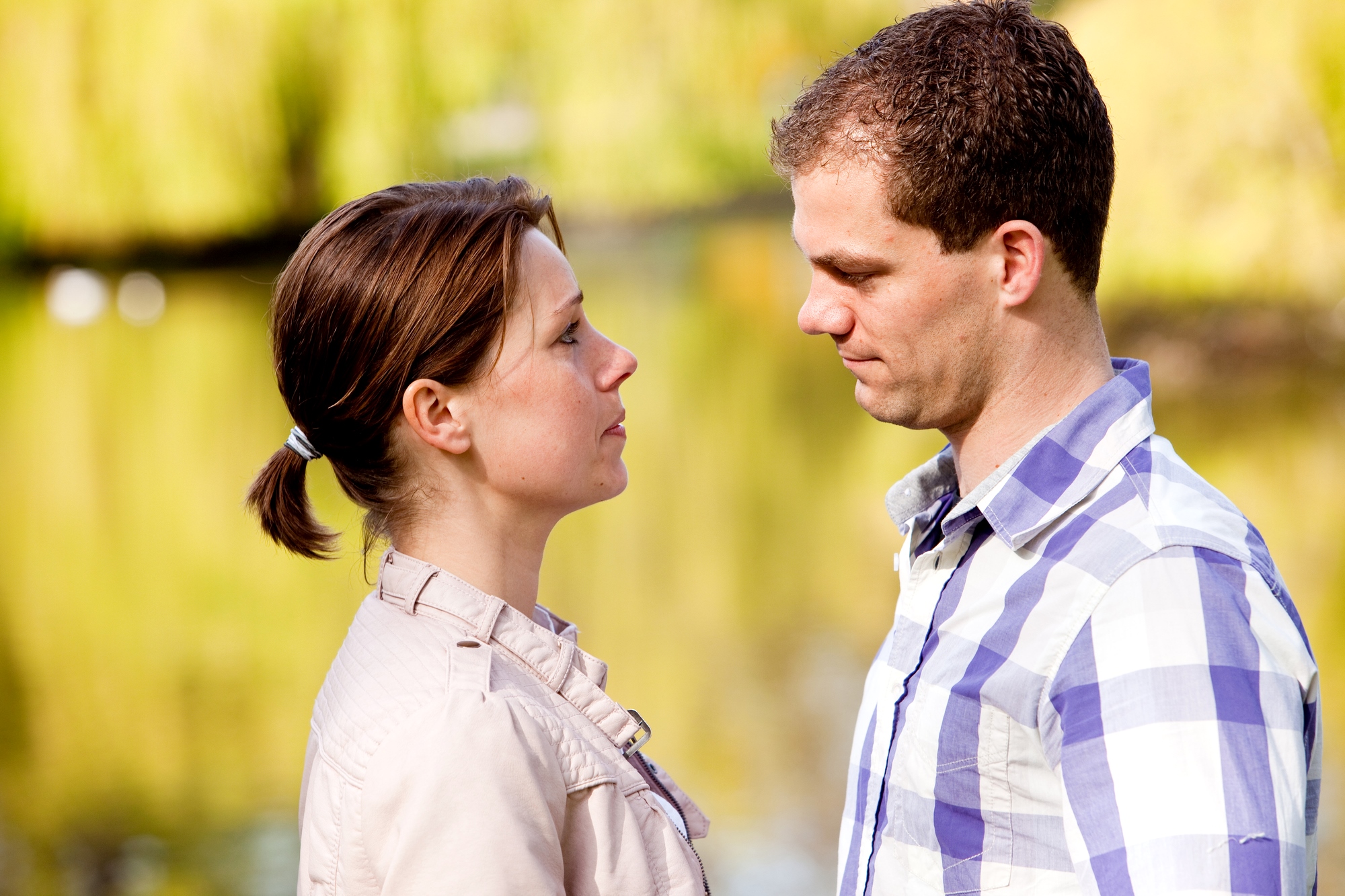 A man and a woman face each other, appearing to be in a serious conversation. They are outdoors near a body of water with greenery in the background. The woman has her hair tied back and wears a light jacket, while the man has short hair and wears a plaid shirt.