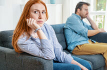 A woman with red hair sits on a couch, resting her chin on her hand, looking thoughtful and slightly distressed. A man with greying hair sits beside her, turned away and looking out the window. Both appear to be in a tense or reflective mood.