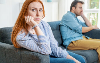 A woman with red hair sits on a couch, resting her chin on her hand, looking thoughtful and slightly distressed. A man with greying hair sits beside her, turned away and looking out the window. Both appear to be in a tense or reflective mood.