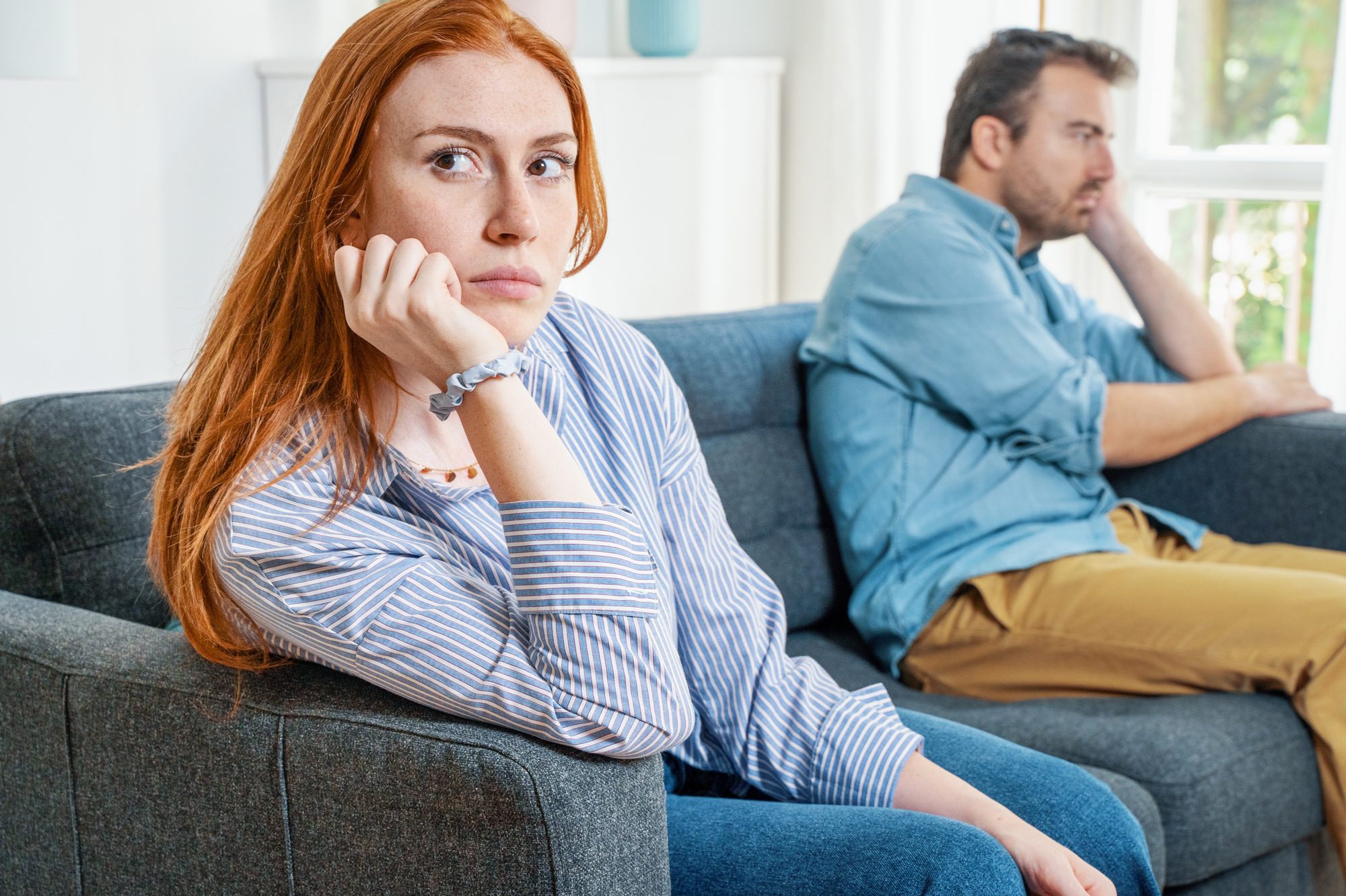 A woman with red hair sits on a couch, resting her chin on her hand, looking thoughtful and slightly distressed. A man with greying hair sits beside her, turned away and looking out the window. Both appear to be in a tense or reflective mood.