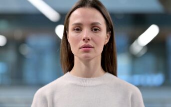 A woman with long brown hair, wearing a light-colored sweater, stands facing the camera with a neutral expression. The background is blurred, featuring a softly lit indoor environment with indistinct light fixtures.