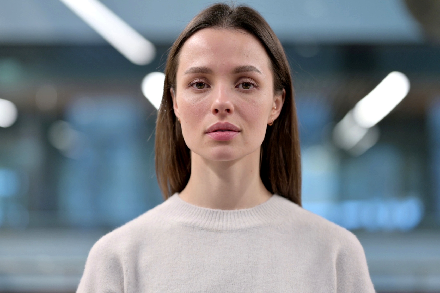 A woman with long brown hair, wearing a light-colored sweater, stands facing the camera with a neutral expression. The background is blurred, featuring a softly lit indoor environment with indistinct light fixtures.