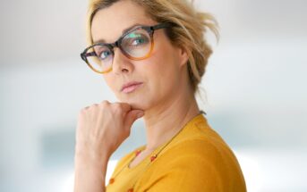 A woman with blond hair and wearing glasses stares intently at the camera, resting her chin on her hand. She is dressed in a mustard yellow top and has a thoughtful expression. The background is out of focus, suggesting an indoor setting.