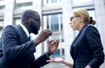 A man and woman, both in business attire and wearing glasses, are engaged in an intense conversation outside a modern office building. The man gestures emphatically with his hands, while the woman looks at him with a serious expression, holding one hand open.