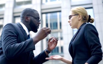 A man and woman, both in business attire and wearing glasses, are engaged in an intense conversation outside a modern office building. The man gestures emphatically with his hands, while the woman looks at him with a serious expression, holding one hand open.