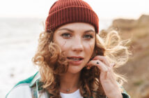 A person with curly hair wearing a red beanie and a green jacket is standing outdoors near a cliff and the ocean. They are looking directly at the camera, with one hand touching their hair. The background is slightly blurred, showing waves and rocky terrain.