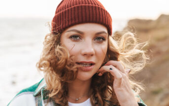 A person with curly hair wearing a red beanie and a green jacket is standing outdoors near a cliff and the ocean. They are looking directly at the camera, with one hand touching their hair. The background is slightly blurred, showing waves and rocky terrain.