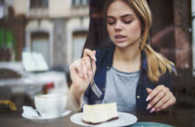 A young woman with blonde hair in a ponytail is sitting at a café table. She is holding a fork and is about to eat a slice of cheesecake on a white plate. There's a cup of coffee with a napkin next to her plate. The background shows a blurred view of buildings.