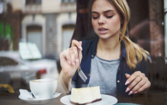 A young woman with blonde hair in a ponytail is sitting at a café table. She is holding a fork and is about to eat a slice of cheesecake on a white plate. There's a cup of coffee with a napkin next to her plate. The background shows a blurred view of buildings.